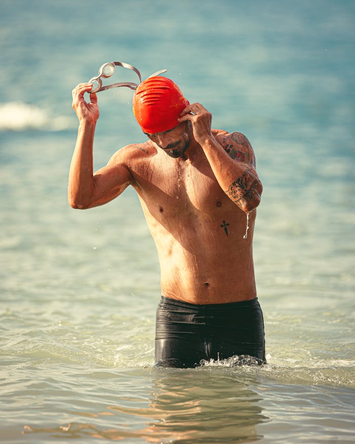 A male swimmer adjusts goggles in the ocean under bright sunlight, wearing a red cap.